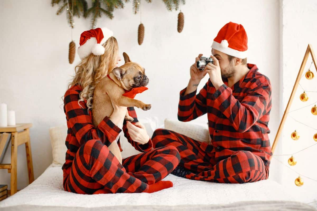 couple in Christmas pajamas, husband is taking a picture of the wife with their dog