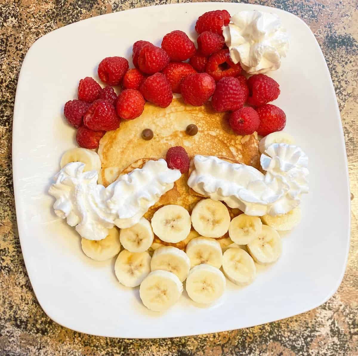 Easy Santa Pancakes for Christmas breakfast with whipped cream for his mustache, bananas for his beard, and raspberries for his hat